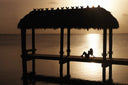 A silhouetted thatched-roof dock structure over calm water at sunset, with two people sitting under the shelter and the sun reflecting on the water surface.