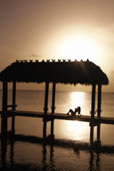 A silhouetted thatched-roof pavilion over calm water at sunset, with a couple sitting on the deck and the sun reflecting on the water surface.