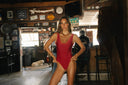 A woman posing in a red one-piece swimsuit inside a rustic bar or restaurant with wooden walls, framed signs, and bar stools in the background.